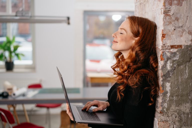 Attractive redhead woman standing daydreaming as she leans against a brick wall in an office holding an open laptop